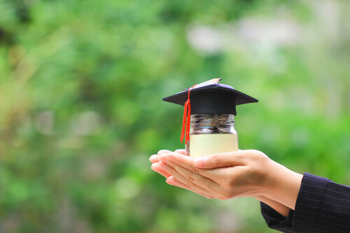 Woman hand holding coins money in glass bottle with graduates hat on natural green background, Saving money for education concept