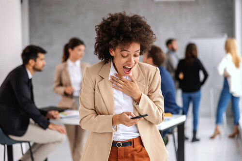 Young excited mixed race businesswoman standing in office and reading message on the phone. Good news. In background are her colleagues working on important project.